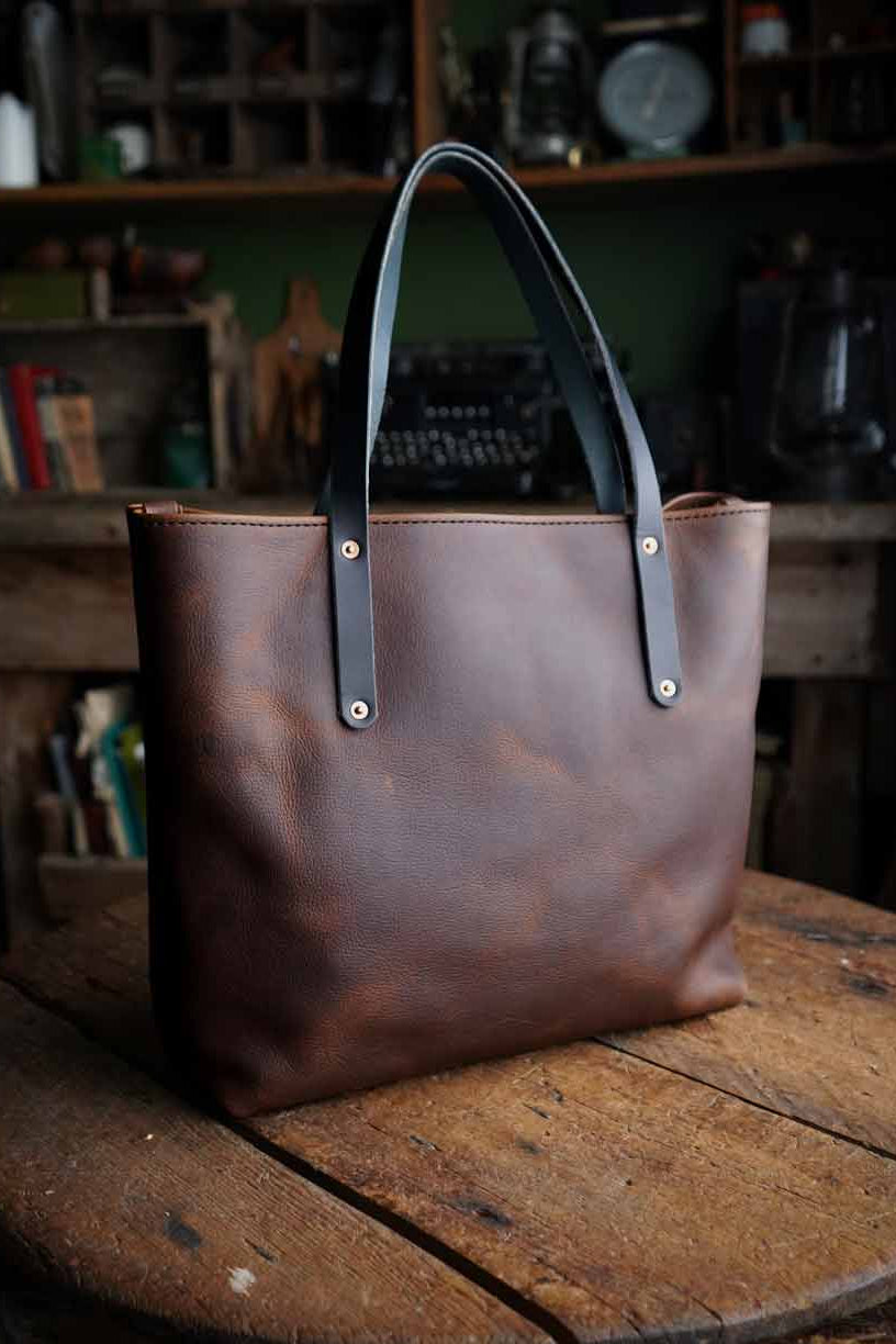 Brown leather tote bag with black handles on a wooden surface in a rustic setting.