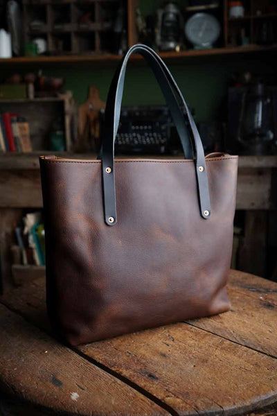 Brown leather tote bag with black handles on a wooden surface in a rustic setting.